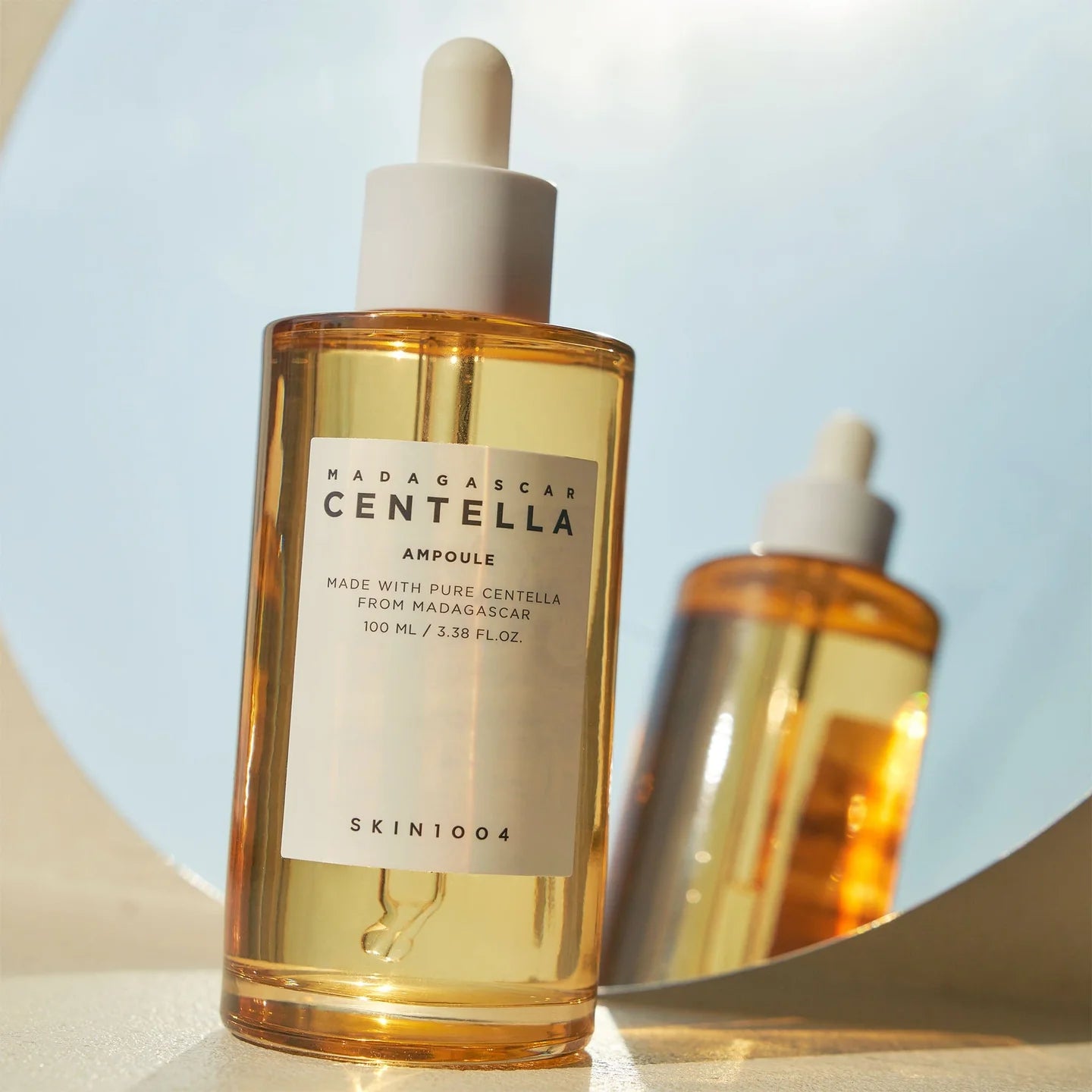SKIN1004 Madagascar Centella Ampoule geplaatst voor een ronde spiegel in warm zonlicht, met spiegeling van het flesje