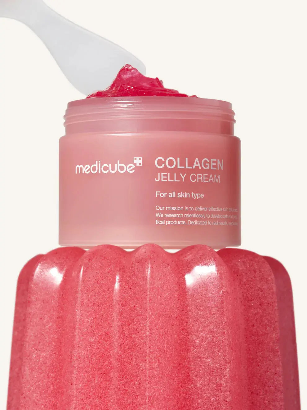 Medicube Collagen Jelly Cream, spatel schept jelly crème uit de pot boven een roze pudding